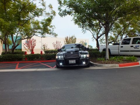 2003 Ford Crown Victoria - Distant front view of lowered black sedan with push bar, halo lights lit, next to white truck.