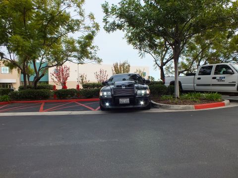 2003 Ford Crown Victoria - Black sedan with custom front guard photographed from across the lot near landscaping and a white truck.