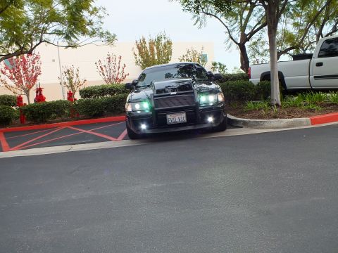 2003 Ford Crown Victoria - Lowered black Crown Victoria front view across lot, custom grille guard, headlights illuminated.