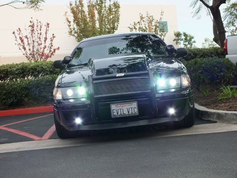 2003 Ford Crown Victoria - Front-on view of lowered black sedan with custom guard and billet grille parked by shrubs.