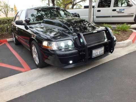 2003 Ford Crown Victoria - Lowered black sedan angled close view, custom grille guard and billet grille, Baker Hughes truck behind.