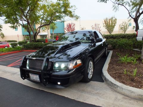 2003 Ford Crown Victoria - Black Ford Crown Victoria parked beside shrubs in a lot, showing brush guard, side profile and chrome wheels.