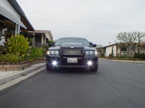 2003 Ford Crown Victoria - Low front angle of a black Crown Victoria with lit fog lamps parked on a street, EVL VIC plate visible.