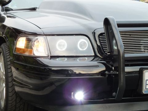 2003 Ford Crown Victoria - Close-up of a modified Crown Victoria's halo projector headlight, billet grille edge and lit fog lamp behind a black push bar.