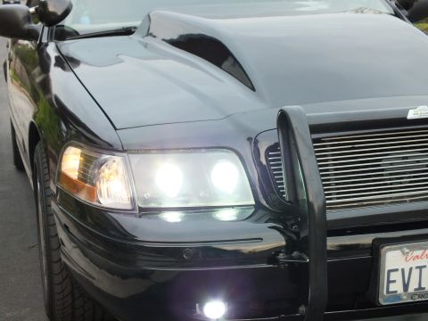 2003 Ford Crown Victoria - Front quarter view of a black Crown Victoria with hood scoop, lit headlights and black push bar.