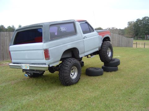 1983 Ford Bronco - Modified Ford Bronco with large tires, elevated on a stack of tires in a grassy yard.