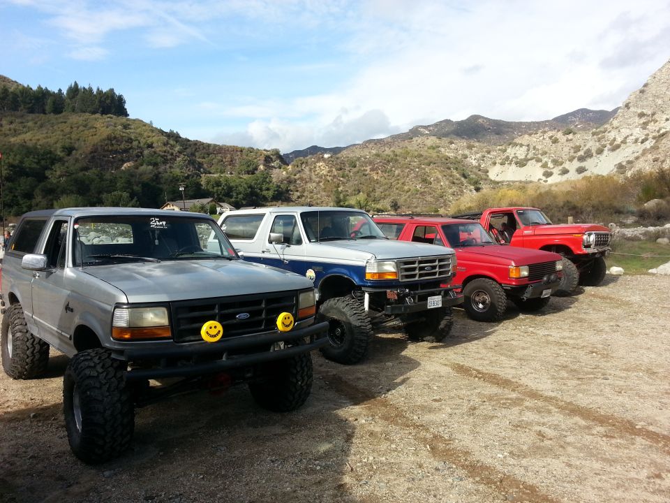 Four off-road vehicles parked in a line on a dirt lot with mountains in the background. The first two are Ford Broncos, followed by two red trucks