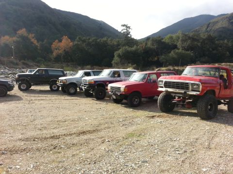 1978 Ford Bronco - A line of modified Ford Broncos and trucks are parked in a dirt lot with mountains in the background.