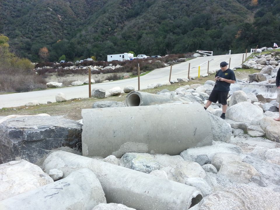Concrete pipes and rocks in a rocky, hilly area with a road and some parked vehicles in the distance