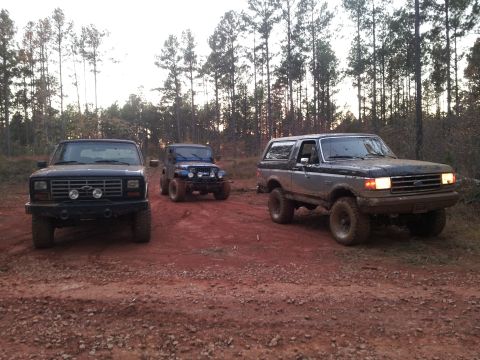 1986 Ford Bronco - Three modified off-road vehicles are parked on a dirt path surrounded by trees.