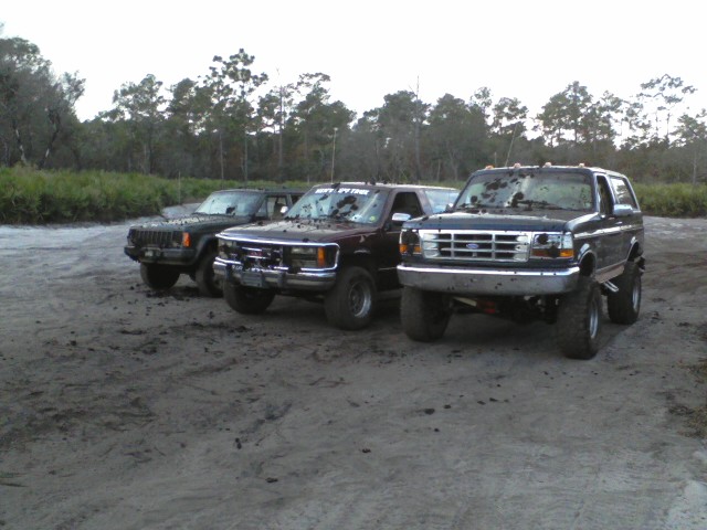 Lifted dark Ford Bronco, maroon pickup, and Jeep Cherokee parked on muddy lot