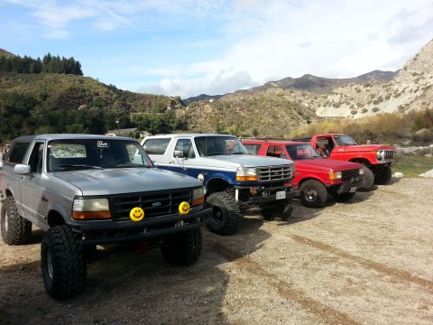 1978 Ford Bronco - A line of modified SUVs and trucks are parked in a dusty, outdoor setting with mountains in the background.