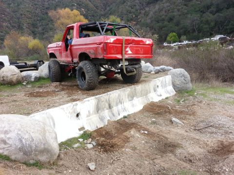 1978 Ford Bronco - A modified red pickup truck navigates an obstacle course with large rocks and concrete barriers.