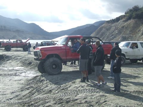 1978 Ford Bronco - A group of people gather around a lifted red 1978 Ford Bronco near a body of water, with other vehicles visible in the background.
