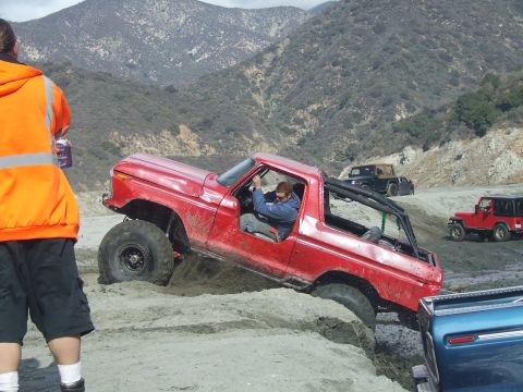 1978 Ford Bronco - A red 1978 Ford Bronco is driven off-road through sand, with other vehicles visible in the background.