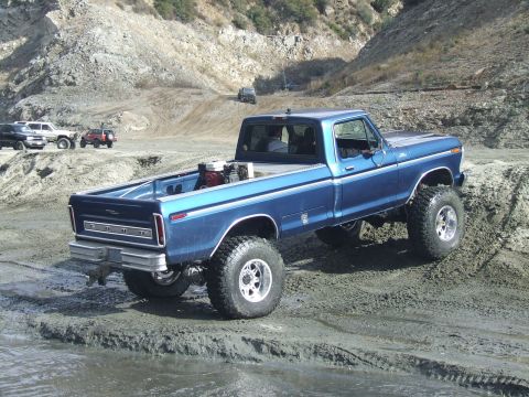 1978 Ford Bronco - Lifted blue Ford pickup truck navigates a muddy off-road course with other vehicles present.