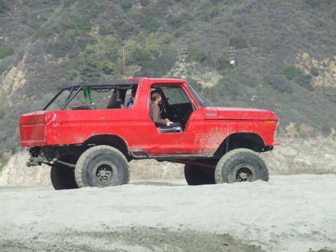 1978 Ford Bronco - A red 1978 Ford Bronco with a roll cage is partially submerged in sand, with a person visible in the driver's seat.