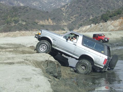 1978 Ford Bronco - A silver Ford Bronco is shown tackling a challenging off-road trail, with another vehicle visible in the distance.