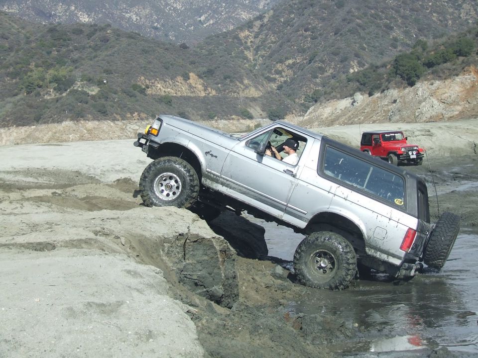 Silver Ford Bronco navigating a muddy incline with a red Jeep in the background