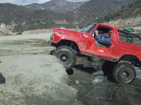 1978 Ford Bronco - A red 1978 Ford Bronco is shown off-roading through a muddy canyon with a driver inside.