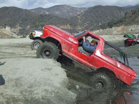 1978 Ford Bronco - A red off-road truck navigates a muddy terrain with a silver truck following closely behind.