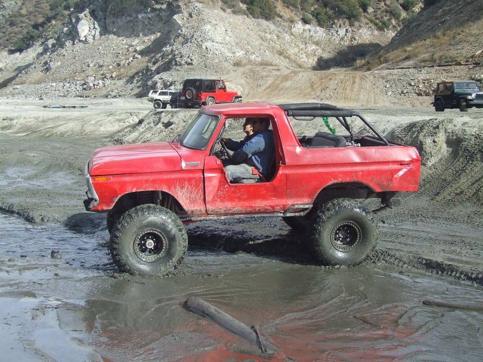 Red 1978 Ford Bronco with large tires driving through mud, roll cage visible