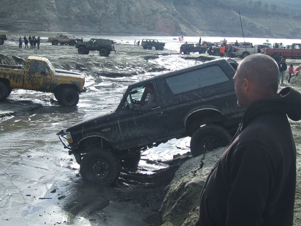 Black Ford Bronco driving through mud with other off-road vehicles in background