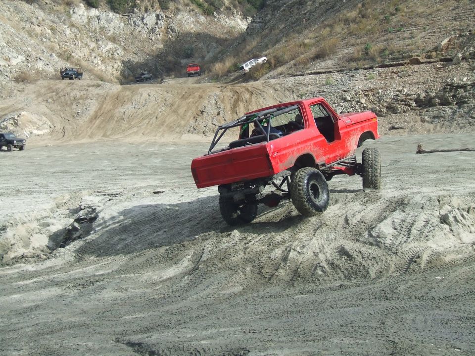 Red 1978 Ford Bronco with roll cage climbing a dirt hill, lifted suspension, off-road tires