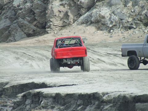 1978 Ford Bronco - A red off-road truck with a roll cage drives on a dirt track, with another truck visible to the right.