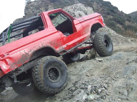 1978 Ford Bronco - A red 1978 Ford Bronco is shown off-roading on a rocky, uneven surface, demonstrating its modified suspension and tires.