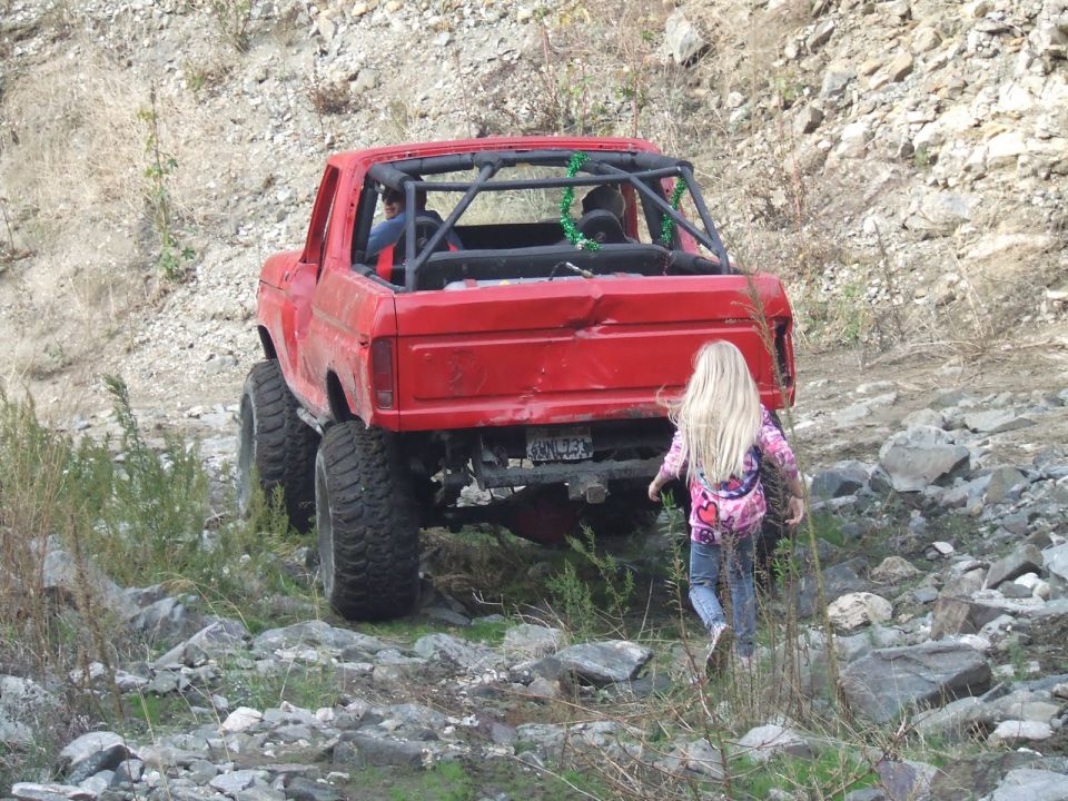 Red 1978 Ford Bronco with a roll cage and large tires, seen from the rear in a rocky, outdoor environment