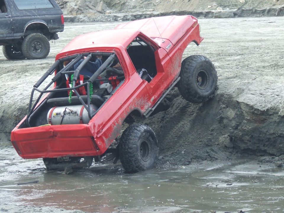 Red Bronco with roll cage going up a muddy hill, another Bronco behind it