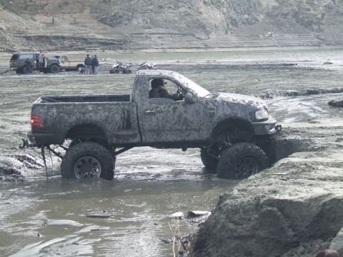 1978 Ford Bronco - A lifted pickup truck navigates a muddy terrain, with other vehicles and people visible in the background.