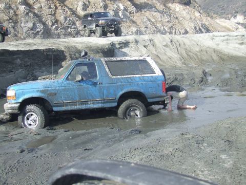 1978 Ford Bronco - A blue Ford Bronco is stuck in a muddy ditch while a person inspects the rear, with another vehicle visible on the embankment above.