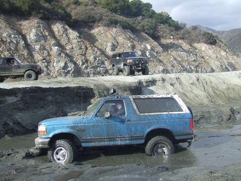 1978 Ford Bronco - Multiple off-road vehicles navigate a muddy terrain, with one blue Ford Bronco partially submerged.