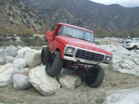 1978 Ford Bronco - A red pickup truck navigates a rocky terrain, with other vehicles in the background.