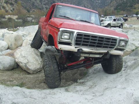 1978 Ford Bronco - This red pickup truck is shown off-roading over large rocks, with other vehicles in the background.