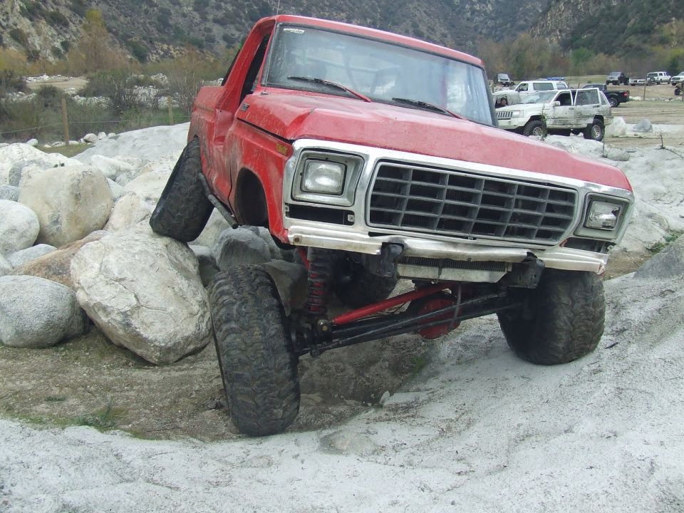 Red pickup truck with lifted suspension navigating large rocks. Other vehicles visible in background