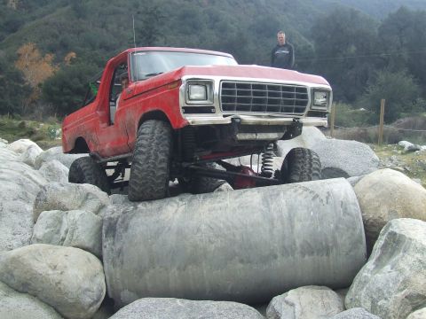 1978 Ford Bronco - A modified red truck navigates a rocky terrain with a person standing in the background.