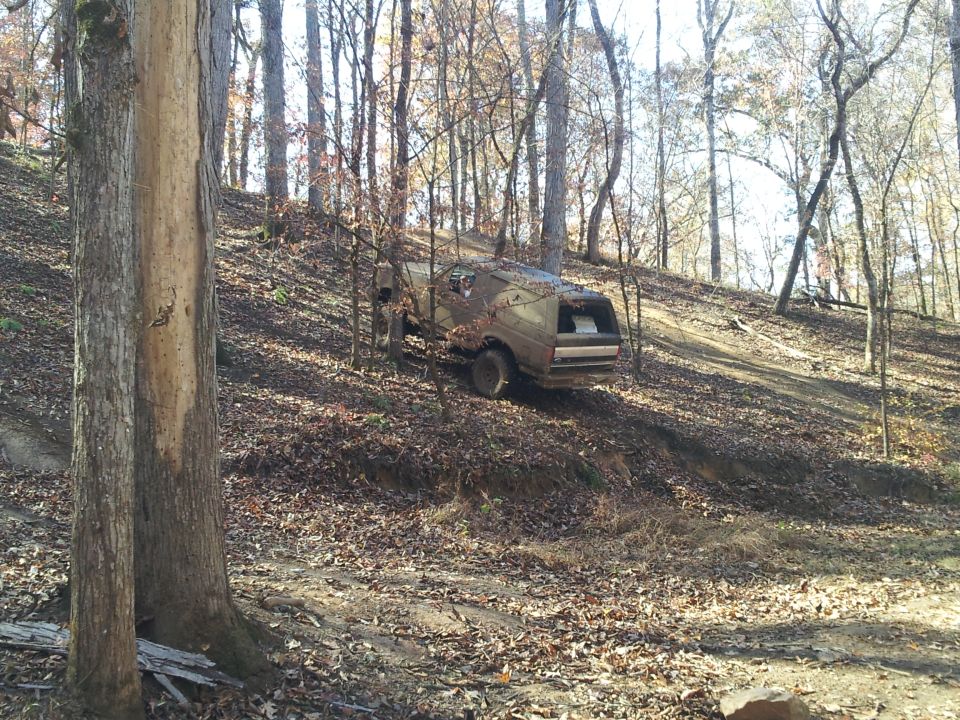Brown SUV with a cap driving up a dirt hill in a forest. Appears to be a 1980s model