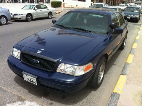 2007 Ford Crown Victoria - A dark blue police car is parked on the side of a street with other vehicles in the background.
