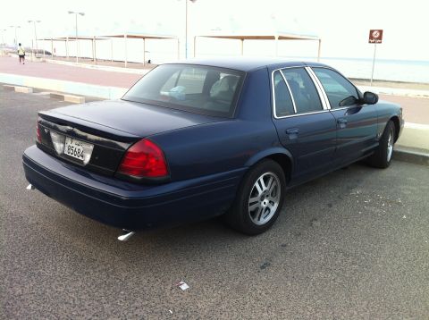2007 Ford Crown Victoria - A dark blue 2007 Ford Crown Victoria is seen from the rear, parked near a waterfront area with a promenade and a pedestrian walkway.