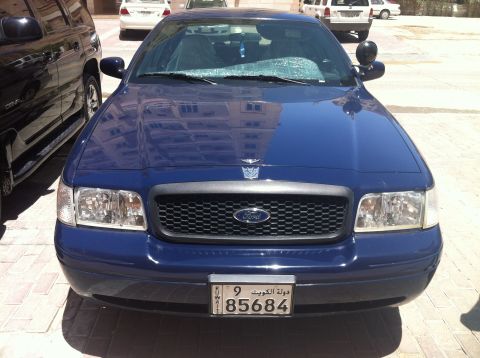 2007 Ford Crown Victoria - A blue 2007 Ford Crown Victoria is parked in front of a building, with a black SUV to its left.