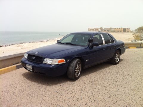 2007 Ford Crown Victoria - A dark blue 2007 Ford Crown Victoria is parked on a sandy lot next to a beach with buildings in the background.