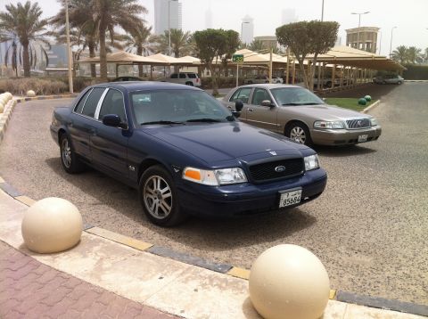 2007 Ford Crown Victoria - Two sedans are parked on a street with palm trees and modern buildings in the background.