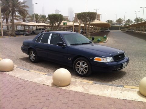 2007 Ford Crown Victoria - A dark blue 2007 Ford Crown Victoria is seen parked in a lot with palm trees and modern buildings in the background.