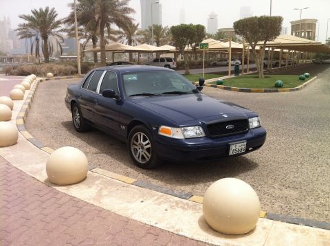 2007 Ford Crown Victoria - A dark blue 2007 Ford Crown Victoria is parked curbside, with a cityscape visible in the background.