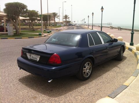 2007 Ford Crown Victoria - A dark blue sedan is parked along a waterfront promenade with a pier in the distance.