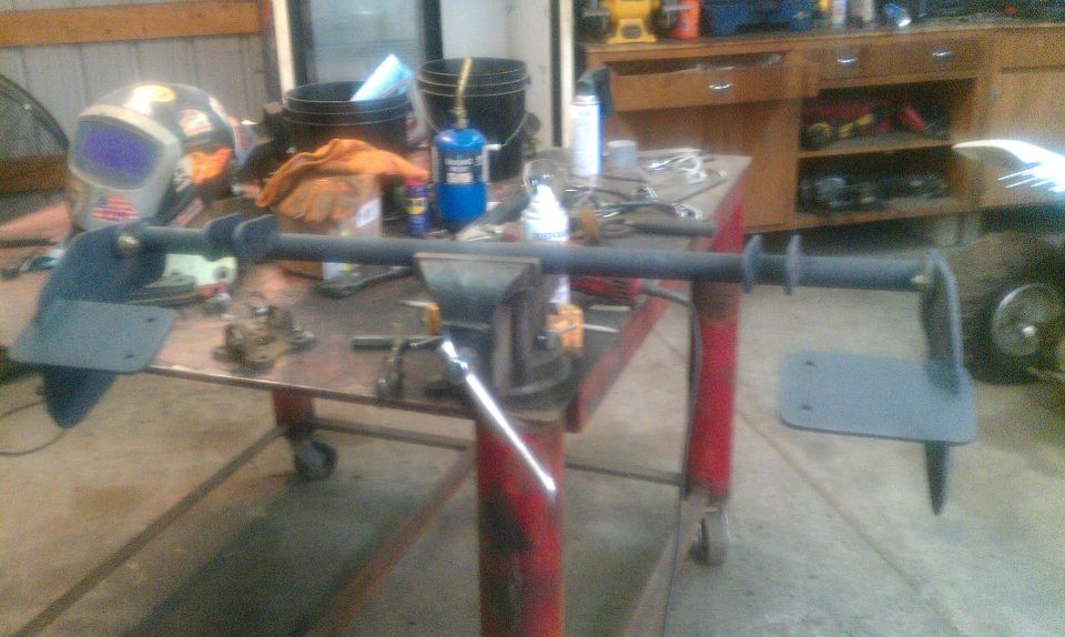 metal bar with brackets clamped in a vise on a workbench, welding mask nearby