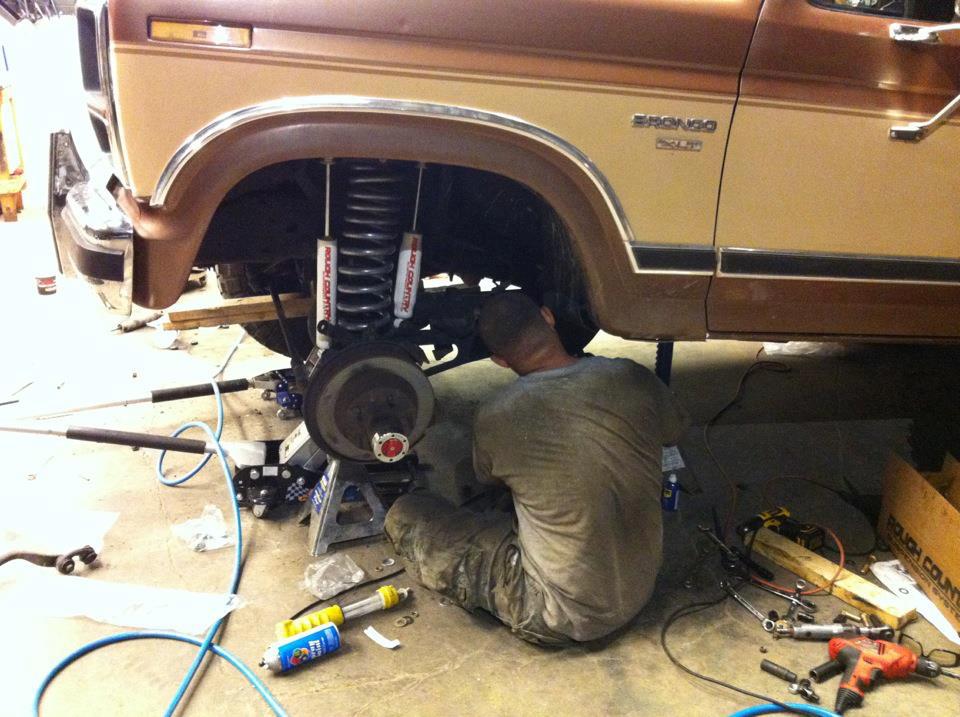 Tan Ford Bronco on jack stand with person working on front suspension in garage